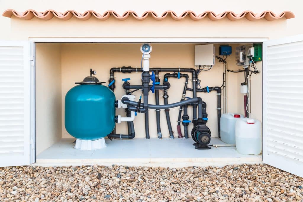 Swimming pool equipment system with a large filter tank, circulation pump, connected pipes, valves, and chemical containers housed in an outdoor utility enclosure.