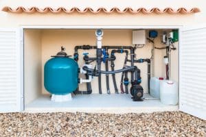 Swimming pool equipment system with a large filter tank, circulation pump, connected pipes, valves, and chemical containers housed in an outdoor utility enclosure.