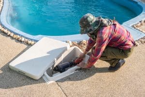 A pool technician repairs a swimming pool pump system in an open equipment compartment beside a residential swimming pool.